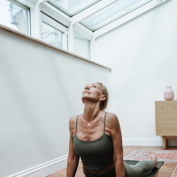 Elderly woman in yoga pose on mat in sunlit room, promoting fitness and wellness.
