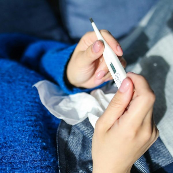 A woman checks a digital thermometer while lying indoors, showing signs of illness.